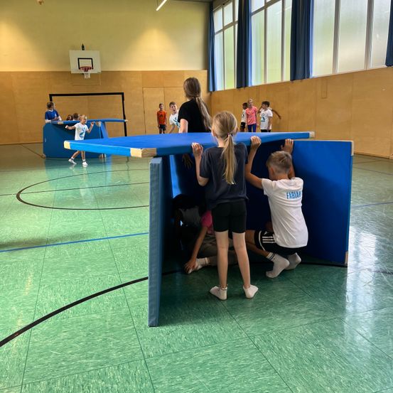 Kinder spielen in einer Turnhalle mit einem blauen Hindernisparcours. Einige krabbeln unter einem Tisch, während andere auf dem Boden spielen.