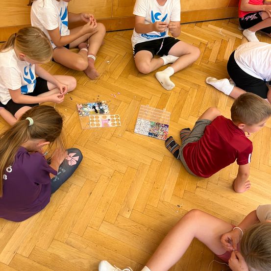Eine Gruppe von Kindern sitzt auf dem Boden und ist in einer Bastelaktivität verwickelt. Sie tragen Freizeitkleidung und scheinen gemeinsam an einem Projekt zu arbeiten.