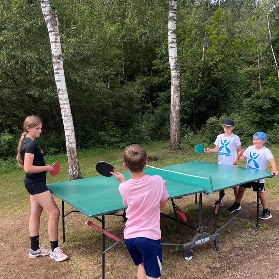 Vier Kinder spielen Tischtennis auf einem grünen Tisch im Freien, mit Bäumen und Gras im Hintergrund.