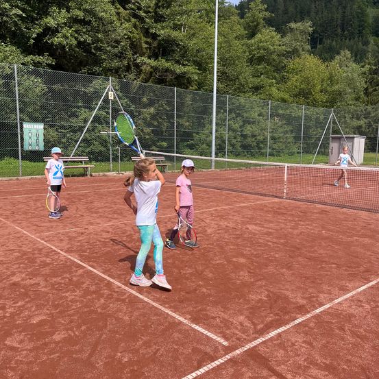 Vier Kinder spielen Tennis auf einem Sandplatz. Ein Kind hält einen Tennisschläger, während ein anderes hinter dem Netz steht. Der Platz ist von einem Zaun und Bäumen umgeben.