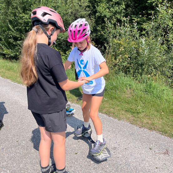 Zwei junge Mädchen stehen auf einem Skateboard, eines trägt einen Helm und das andere hält ein Skateboard. Sie sind auf einem asphaltierten Weg mit Gras und Bäumen im Hintergrund.