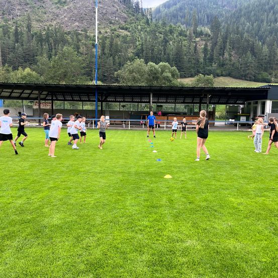 Eine Gruppe von Kindern spielt ein Spiel auf einem Rasenfeld mit einem Fußball und Hütchen, unter einem überdachten Pavillon mit Bergen im Hintergrund.
