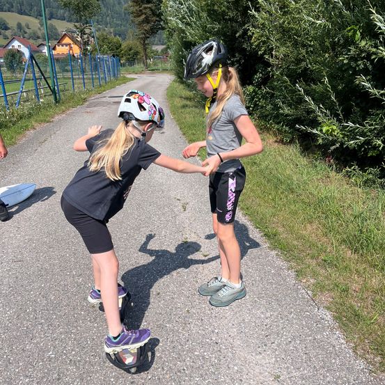 Zwei Mädchen stehen auf Einrädern auf einem Schotterweg, eine hält die Hand der anderen. Sie tragen beide Helme und Turnschuhe.
