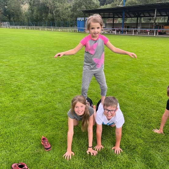 Drei Kinder spielen auf einem grünen Feld. Ein Mädchen steht auf dem Rücken zweier anderer Kinder, die Liegestütze machen.