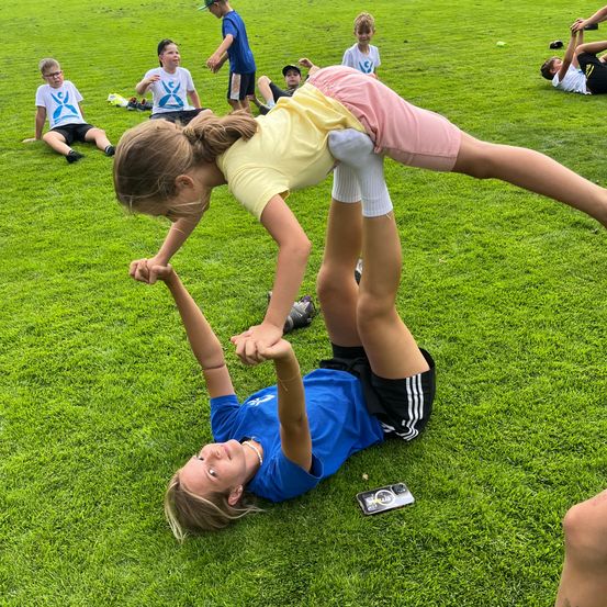 Eine Gruppe von Kindern spielt auf einem Rasenfeld, ein Mädchen liegt auf dem Boden, während ein anderes Mädchen auf ihren Händen über ihr steht.