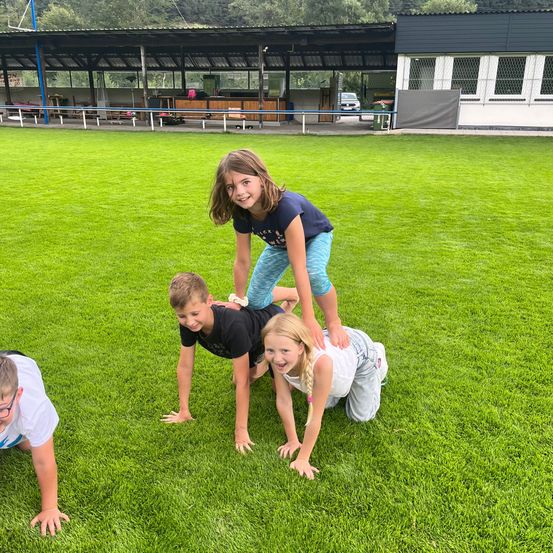 Drei Kinder spielen auf einem grünen Feld. Ein Mädchen steht auf den Schultern von zwei anderen. Sie alle lächeln. Hinter ihnen steht ein Gebäude mit einem geparkten Auto davor.