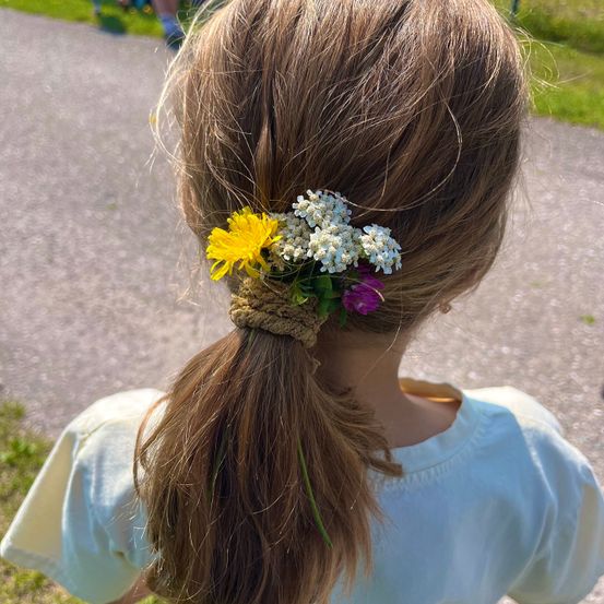 Ein Mädchen mit langen braunen Haaren hat einen Pferdeschwanz, der mit einer Mischung aus gelben und weißen Blumen geschmückt ist. Sie steht auf einem Weg.