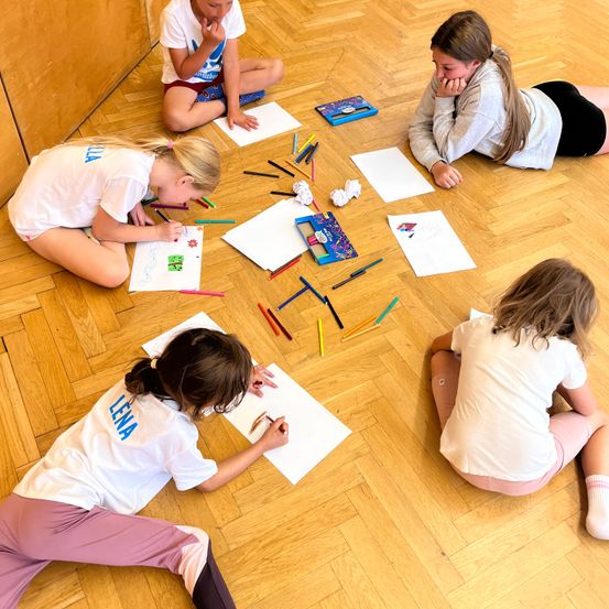 Fünf junge Mädchen sitzen auf einem Holzboden und sind mit einer Malaktivität beschäftigt. Jedes Mädchen hat ein Stück Papier und einen Satz Buntstifte. Einige konzentrieren sich auf ihre Zeichnungen, während andere umherschauen. Der Raum hat einen Holzboden und eine Holzwand im Hintergrund.