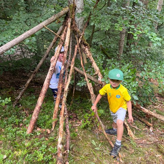 Zwei junge Jungen bauen in einem Wald eine Unterkunft aus Stöcken. Ein Junge ist im Inneren der Unterkunft, der andere draußen.