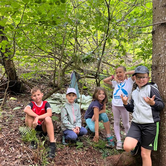 Fünf Kinder posieren für ein Foto im Wald, wobei eines gegen einen Baumstamm gelehnt ist. Sie alle lächeln und scheinen ihre Zeit im Freien zu genießen.