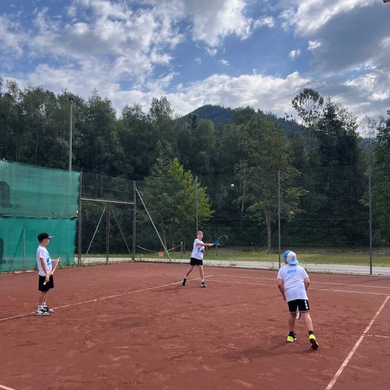 Drei Kinder spielen Tennis auf einem Sandplatz. Ein Kind schlägt den Ball, während die anderen beiden stehen und zusehen. Der Platz ist von einem Zaun und Bäumen umgeben.