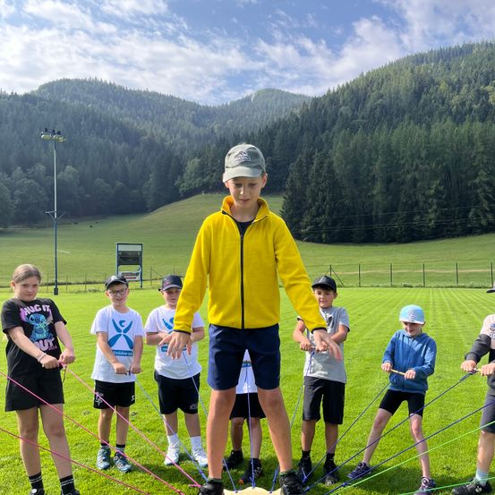Eine Gruppe von Kindern auf einem Rasenfeld, ein Kind in einer gelben Jacke steht auf einem Balancebrett und hält Seile, während andere Seile um ihn herum halten.