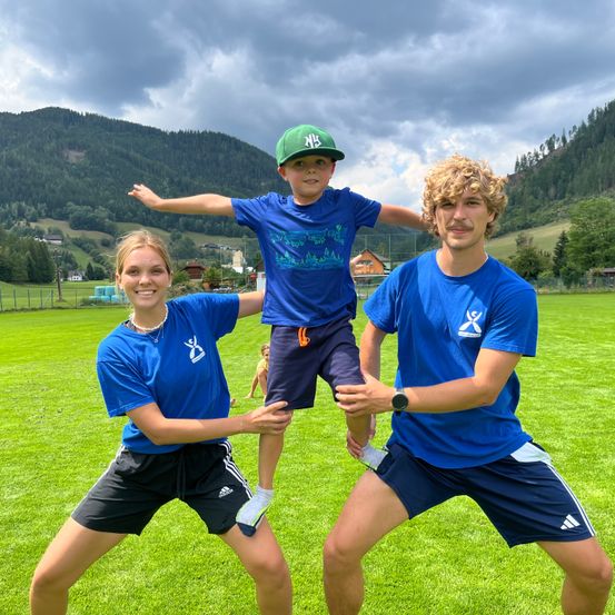 Drei Personen in blauen Shirts halten einen kleinen Jungen in der Luft auf einem Rasenfeld mit Bergen im Hintergrund.