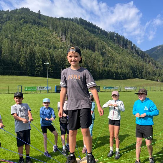 Eine Gruppe von Kindern steht auf einem Rasenfeld und hält an Seilen fest, wobei ein Junge in der Mitte ein Puma-Shirt und schwarze Shorts trägt.