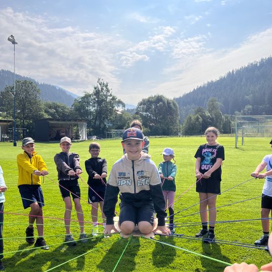 Eine Gruppe von Kindern spielt ein Spiel mit Seilen auf einem Rasenfeld, mit Bergen im Hintergrund.