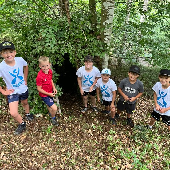 Sechs Jungen stehen in einem Wald und posieren für ein Foto in der Nähe einer selbstgebauten Unterkunft. Sie tragen T-Shirts und Shorts in verschiedenen Farben.
