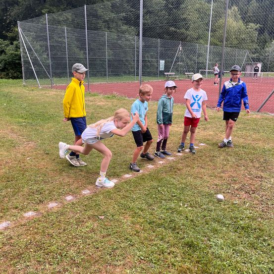 Eine Gruppe von Kindern spielt ein Spiel auf einem Grasfeld mit einem Gitterzaun im Hintergrund. Ein Mädchen in einem weißen Shirt beugt sich herunter, um einen Ball zu treten.