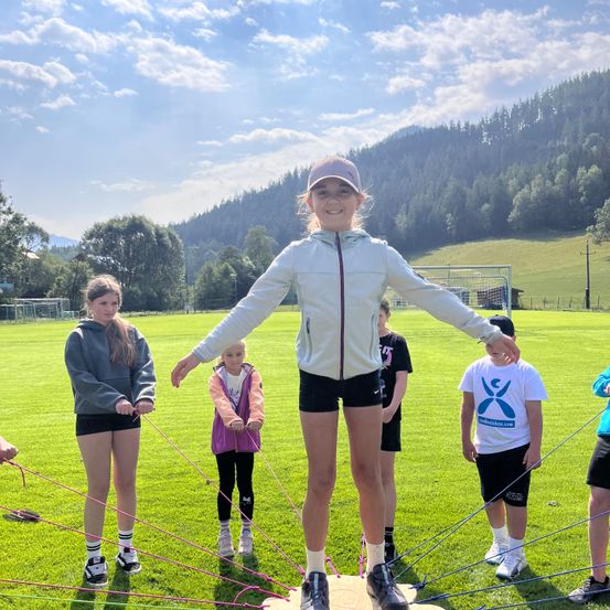Eine Gruppe von Kindern befindet sich auf einem Rasenfeld, wobei ein Mädchen auf einem Balancebrett steht, während andere Seile halten. Das Mädchen trägt eine weiße Jacke und eine Mütze. Hinter ihr hält ein junges Mädchen in einer rosa Jacke ein Seil. Im Hintergrund befindet sich ein Berg mit Bäumen.