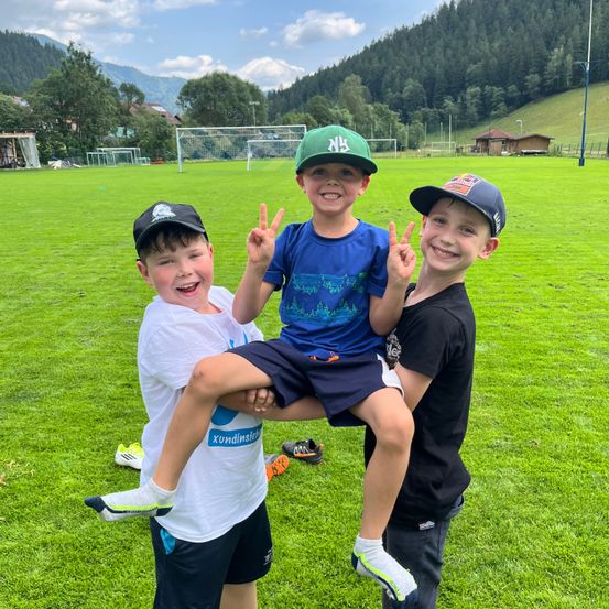 Drei Jungen in Mützen posieren für ein Foto auf einem Rasenfeld mit einem Fußballtor im Hintergrund.