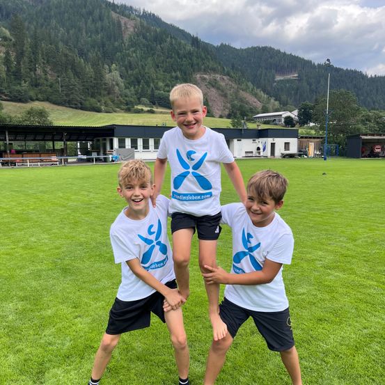 Drei Jungen in identischen weißen T-Shirts mit blauen Logos stehen auf einem Rasenfeld mit Bergkulisse. Ein Junge wird von den anderen beiden hochgehoben.