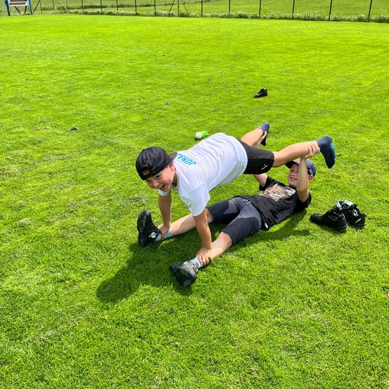 Zwei Jungen liegen auf einem grünen Rasen, einer macht einen Handstand, während der andere auf dem Rücken liegt. Sie tragen beide Turnschuhe und Mützen.