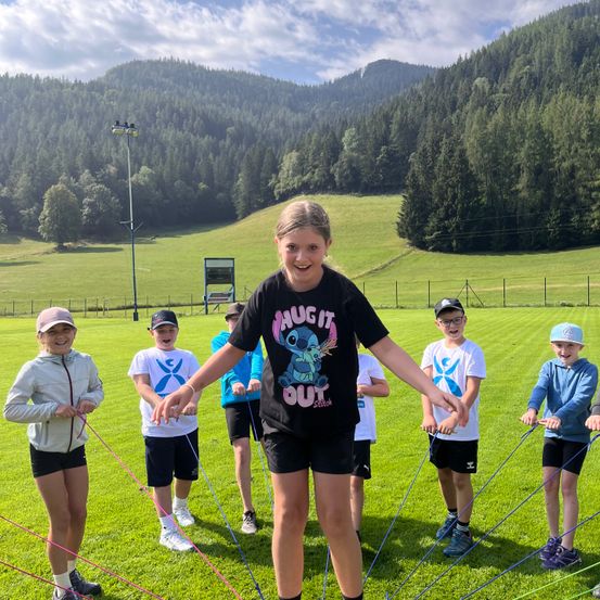 Eine Gruppe von Kindern springt Seil auf einem Rasenfeld mit Bergen im Hintergrund.