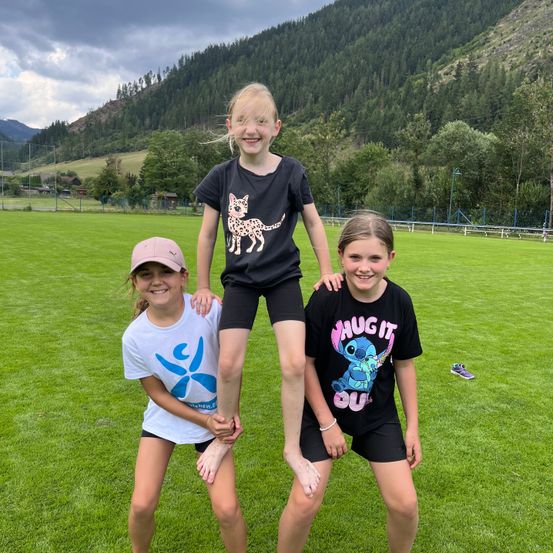Drei Mädchen stehen auf einem Rasenfeld mit einem Berg im Hintergrund. Das Mädchen in der Mitte trägt ein T-Shirt mit Gepardenmuster, während die anderen weiße und schwarze T-Shirts tragen. Sie lächeln und scheinen für ein Foto zu posieren.