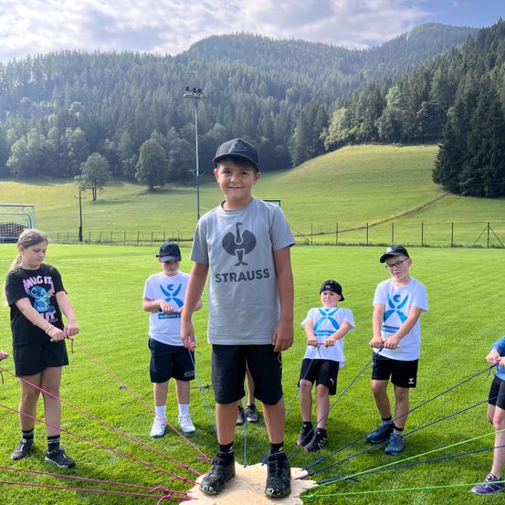 Eine Gruppe von Kindern macht eine Übung mit Widerstandsbändern auf einem Rasenfeld mit Bergen im Hintergrund.
