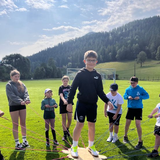 Eine Gruppe von Kindern und Jugendlichen ist in einer Teambuilding-Übung auf einem Rasenfeld mit Bergen im Hintergrund. Sie halten Seile und bilden einen Kreis um einen Jungen in der Mitte.