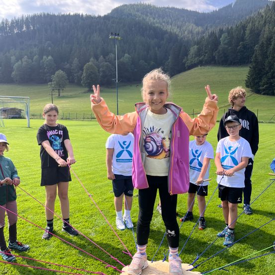 Eine Gruppe von Kindern spielt mit Seilen auf einer Wiese mit Bergen im Hintergrund.