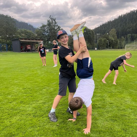Zwei Jungen machen einen Handstand auf einem grünen Feld, während andere Kinder im Hintergrund stehen.