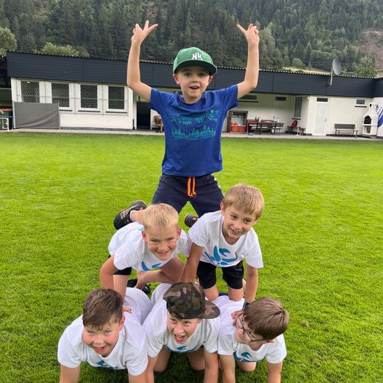 Eine Gruppe junger Jungen bildet eine menschliche Pyramide auf einem grünen Feld mit einem Gebäude im Hintergrund.