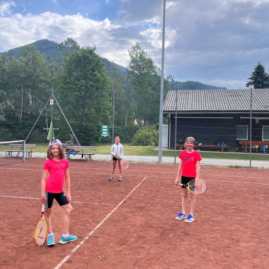 Drei Kinder spielen Tennis auf einem Sandplatz, mit Bergen und Bäumen im Hintergrund.
