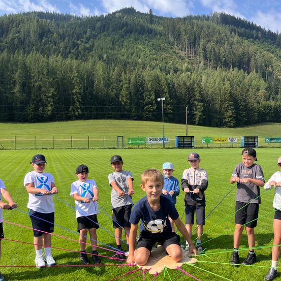 Eine Gruppe von Kindern ist auf einem Rasenfeld mit bunten Seilen bei einer körperlichen Aktivität, umgeben von einem Zaun und einer malerischen Kulisse aus Bäumen und Bergen.