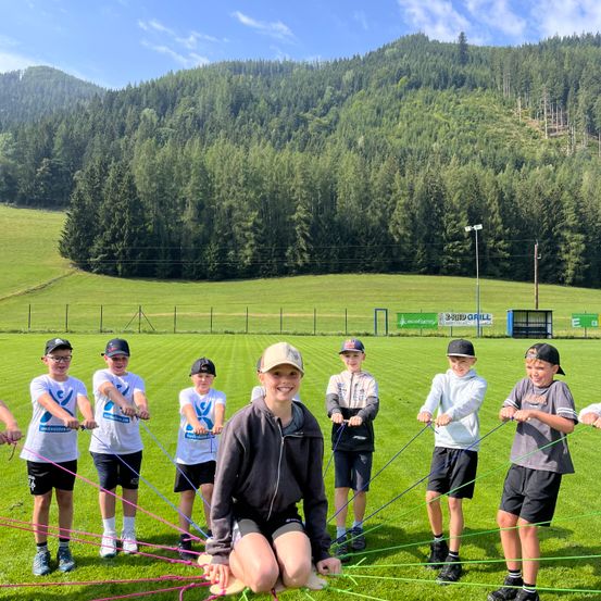 Eine Gruppe von Kindern und einer Frau steht auf einem Rasenfeld und hält bunte Seile. Sie lächeln und scheinen an einer Teambuilding-Aktivität beteiligt zu sein. Hinter ihnen befindet sich ein grünes Feld, ein Zaun und ein Berg mit Bäumen.