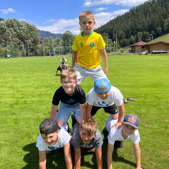 Fünf junge Jungen bilden eine menschliche Pyramide auf einem Rasenfeld mit einem Fußballtor und Häusern im Hintergrund.