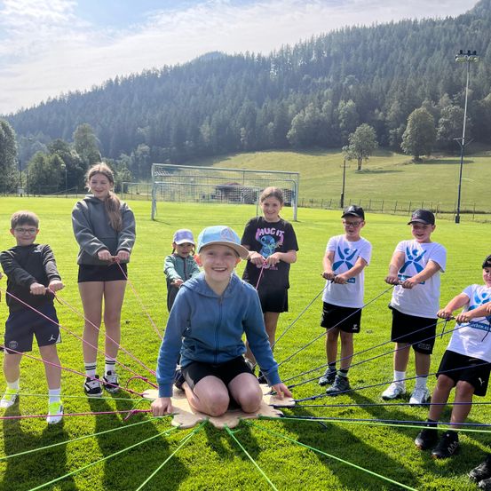 Eine Gruppe von Kindern nimmt an einer Team-Building-Übung auf einem Rasenfeld mit bunten Seilen teil, umgeben von einer malerischen Kulisse aus Bäumen und Bergen.
