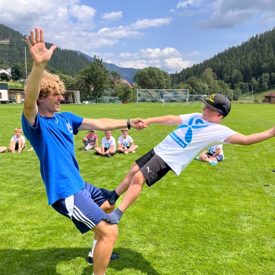 Ein junger Mann in Blau hebt spielerisch ein Kind in einem weißen Shirt mit blauem Logo auf einem Rasenfeld mit Bergen im Hintergrund an.