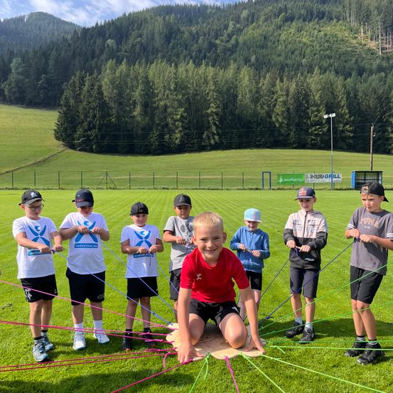 Eine Gruppe junger Jungen hält Seile auf einem Rasenfeld, einer Junge sitzt auf einem Brett. Sie tragen alle Mützen und Turnschuhe. Im Hintergrund befindet sich ein Zaun, ein Berg und ein Banner.