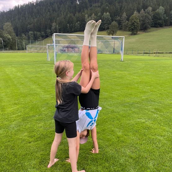 Ein Mädchen steht auf dem Kopf auf einem Rasenfeld, während ein anderes Mädchen ihre Beine hält. Sie befinden sich auf einem Fußballfeld mit einem Tor im Hintergrund.