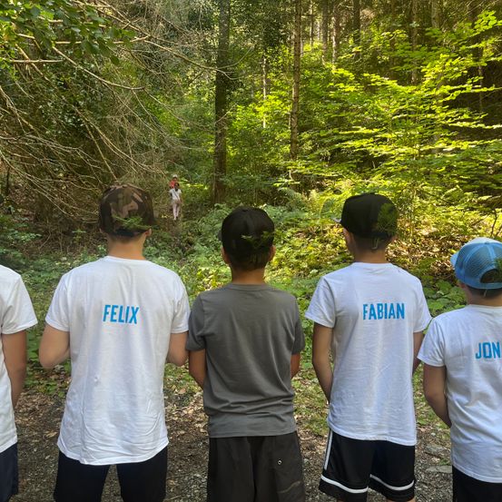 Fünf Jungen in einem Wald, mit dem Rücken zur Kamera. Der Junge auf der linken Seite hat eine Mütze mit einem Tarnmuster. Der Junge neben ihm hat den Namen Felix auf seinem Shirt. Der dritte Junge hat einen schwarzen Hut mit einem grünen Blatt darauf. Der Junge auf der rechten Seite hat den Namen Fabian auf seinem Shirt. Ein weiterer Junge geht in der Ferne.