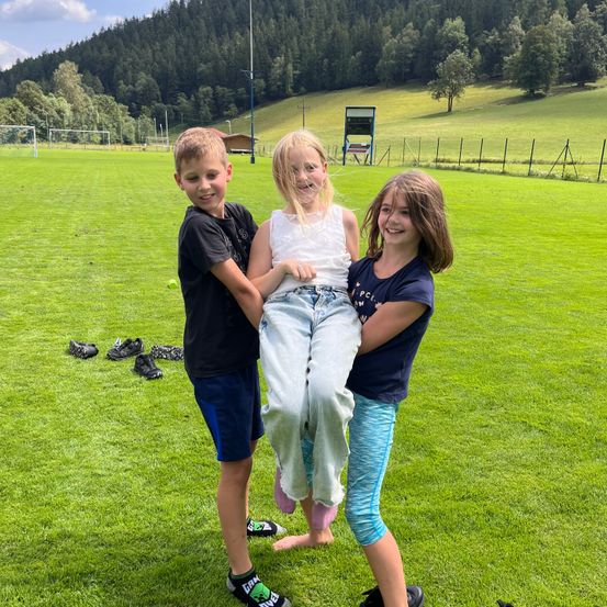 Drei Kinder stehen auf einem Rasenfeld, ein Junge hebt ein Mädchen in die Luft, während ein anderes Mädchen ihre Hand hält.