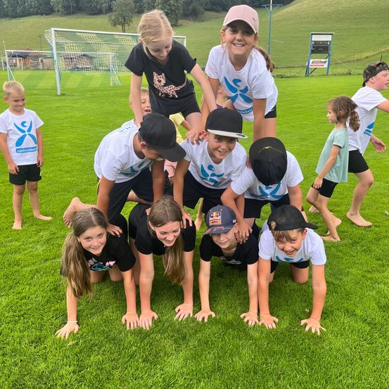 Eine Gruppe von Kindern bildet eine Pyramide auf einem Rasenfeld, mit einem Fußballtor und Häusern im Hintergrund.