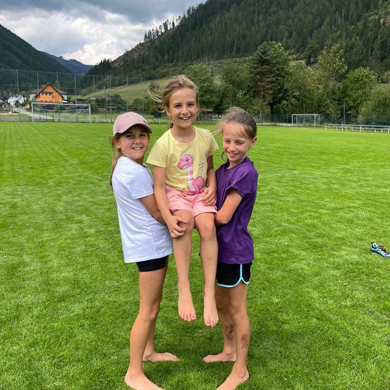 Drei Mädchen stehen auf einem grünen Feld, wobei eines von den anderen hochgehoben wird. Sie alle lächeln und scheinen für ein Foto zu posieren. Hinter ihnen ist ein Rasenfeld mit einem Fußballtor, und in der Ferne sind Berge und Häuser zu sehen.