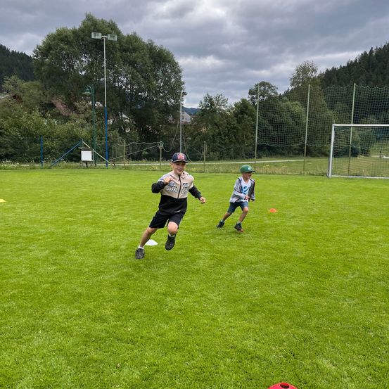Zwei Kinder spielen auf einem Rasenfeld mit orangefarbenen Hütchen und einem Fußballtor im Hintergrund. Ein Kind läuft, während das andere steht.