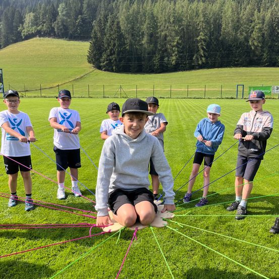 Eine Gruppe von Kindern ist an einer Outdoor-Aktivität auf einem Grasfeld mit bunten Seilen beteiligt, umgeben von einem Zaun und Bäumen.