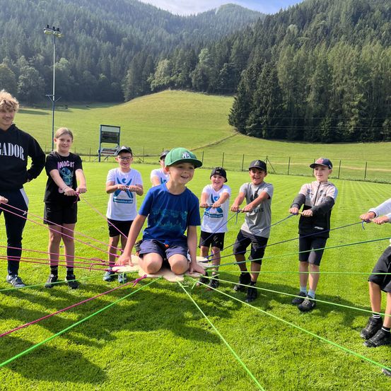 Eine Gruppe von Kindern spielt ein Spiel auf einem Rasenfeld mit bunten Seilen, einige sitzen auf Holzbrettern. Bäume und Berge sind im Hintergrund.