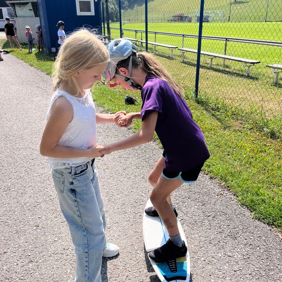 Zwei junge Mädchen halten Händchen; eine steht auf einem Skateboard, während die andere auf dem Boden steht. Dahinter befindet sich ein Grasfeld mit einem Metallzaun und leeren Tribünen.