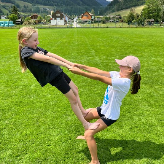 Zwei junge Mädchen spielen auf einem grünen Feld, ein Mädchen wird vom anderen hochgehoben, während sie beide lächeln. Hinter ihnen befinden sich Häuser und ein Zaun. Der Himmel ist klar.