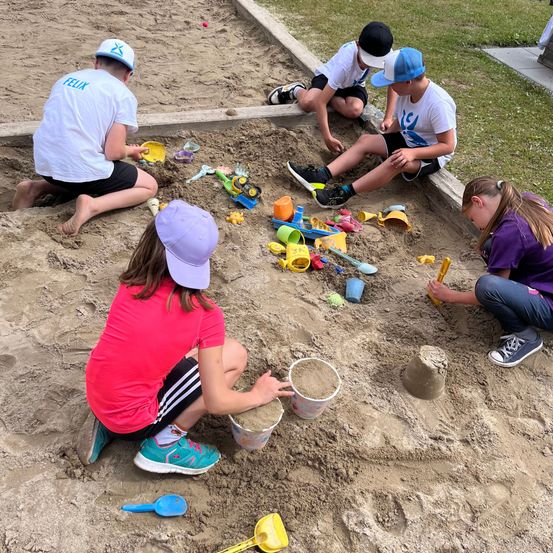Fünf Kinder spielen in einem Sandkasten mit verschiedenen Spielzeugen, einschließlich Eimern und Schaufeln. Sie tragen Mützen und Turnschuhe. Der Sand ist braun.